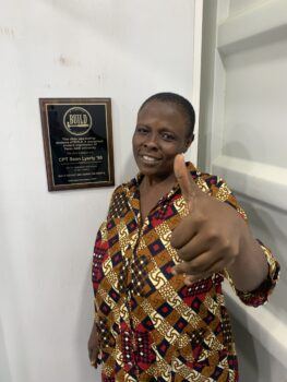 A photo of a woman in a dress giving thumbs up next to a plaque, which states that the clinic she is standing in is dedicated to CPT Sean Lyerly '98