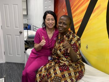A photo of two woman sitting side-by-side in a portable clinic giving thumbs up. One woman is wearing pink scrubs and the other is wearing a dress