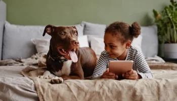 A photo of a young girl in a bed looking at a tablet with a dog next to her on the bed.