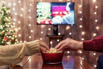 A photo showing two people watching television during the Christmas holidays at home while each is reaching for a bowl of popcorn between them.