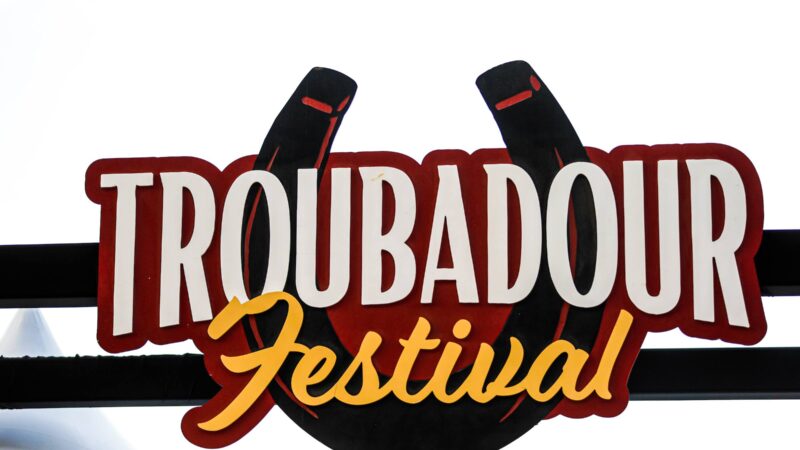 a photo of the Troubadour Festival sign at Aggie Park, with features the name of the event on top of a large red and black horseshoe