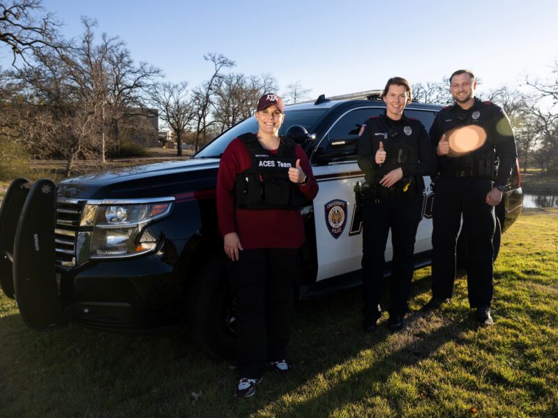 a photo of three people gathered in front of a police SUV giving the thumbs up - one is wearing a maroon shirt and cap along with a vest marked 