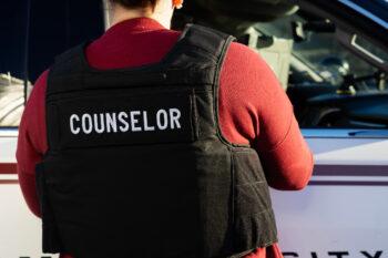 a photo of a woman in a maroon shirt and a black vest labelled "COUNSELOR" standing by a police car with her back to the camera