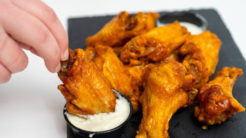 A hand dips a buffalo wing into some dressing with other wings sitting on a plate.