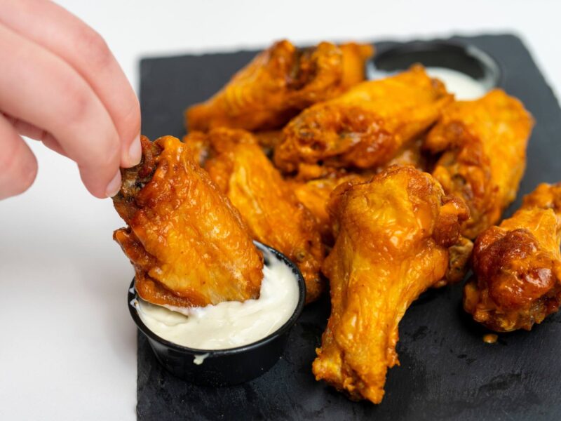 A hand dips a buffalo wing into some dressing with other wings sitting on a plate.