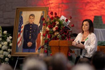A photo of a woman at a lectern speaking at a funeral with a framed painting of a Marine Corps veteran surrounded by flowers to the side of the lectern.