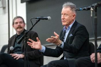A photo of a man speaking in a classroom with a microphone stand in front of him and another man in the background.
