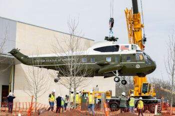 A crane lifts a Marine One helicopter in the air as construction workers in hard hats look on