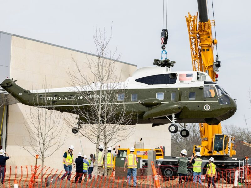 A crane lifts a Marine One helicopter in the air as construction workers in hard hats look on