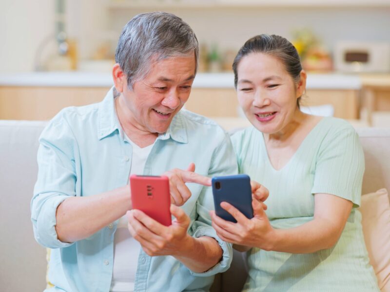 A photo of an Asian elderly couple asian elderly couple looking at their mobile phones together while sitting on a couch.