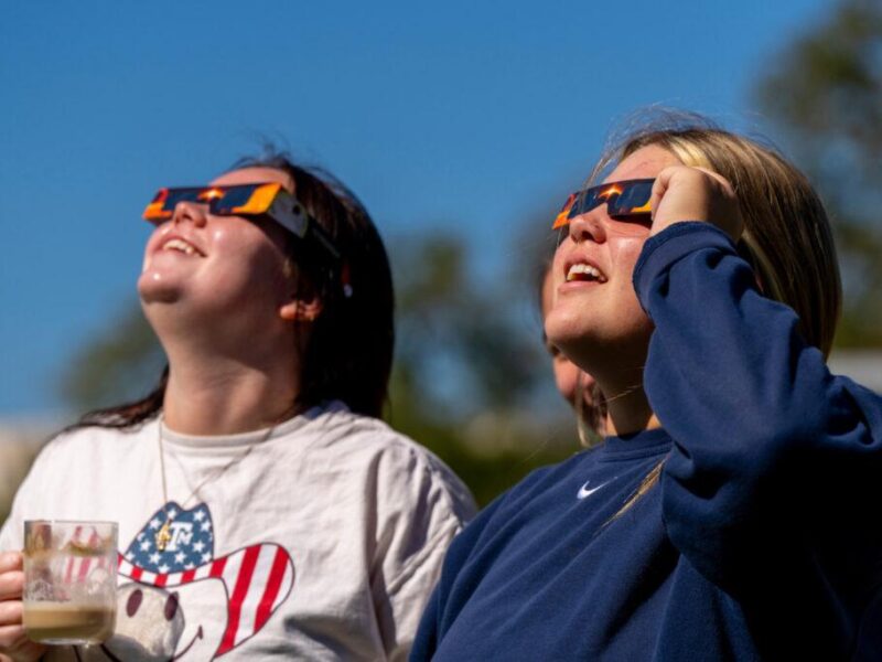Two women wearing eclipse glasses look up at the sky.