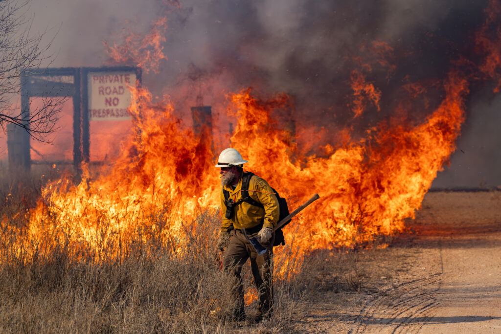 a photo of a man in wildland firefighting gear walking along a dirt road with flames behind him