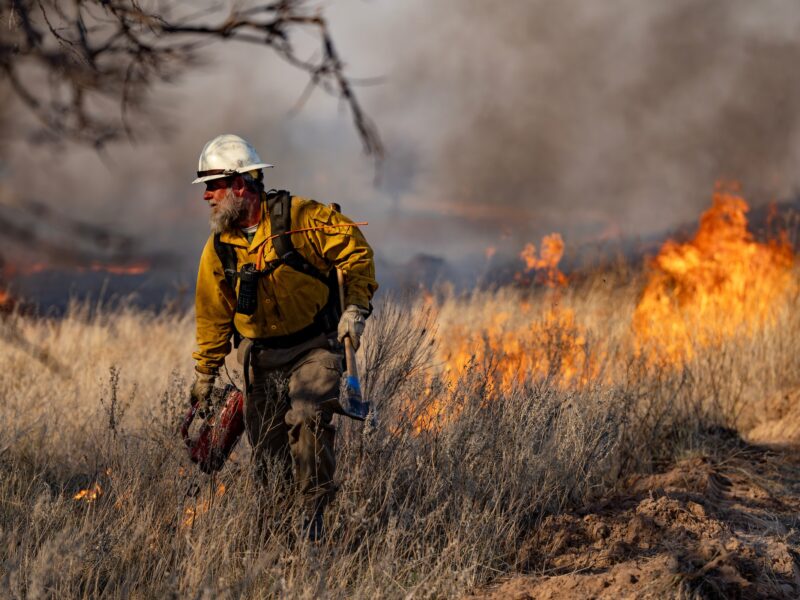 A photo of a man in wildland firefighting gear walking through brush with flames and dark smoke behind him