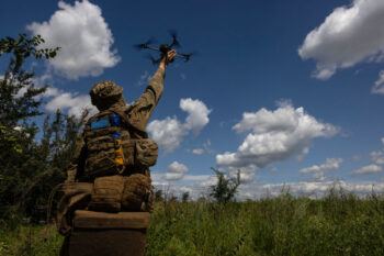a photo of a man in full camouflage holding a quadcopter drone in a grassy field