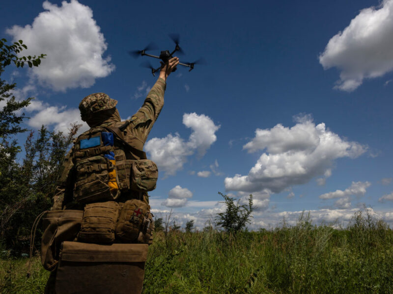 a photo of a man in full camouflage holding a quadcopter drone in a grassy field