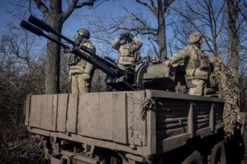 A photo of three people in military uniforms standing on the back of a truck next to a large anti-aircraft gun. One is looking up at the sky through binoculars.