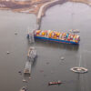 An aerial view of a large cargo ship stacked with shipping containers after running into a bridge. The bridge is collapsed into the water.