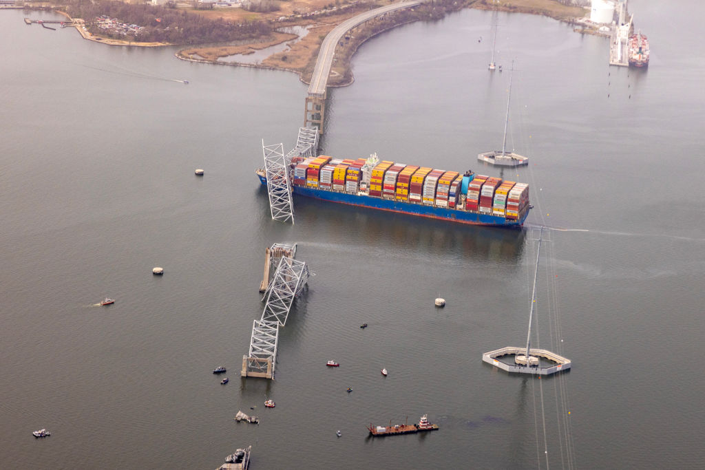 An aerial view of a large cargo ship stacked with shipping containers after running into a bridge. The bridge is collapsed into the water.