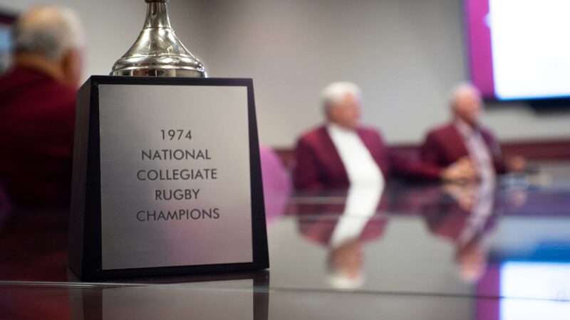 the 1974 national collegiate rugby champion trophy sitting on a table with team members in the background
