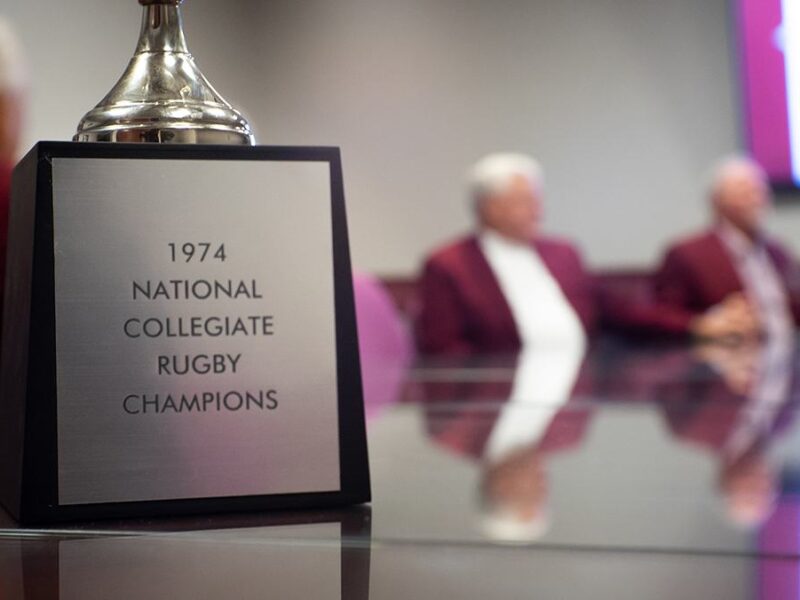the 1974 national collegiate rugby champion trophy sitting on a table with team members in the background
