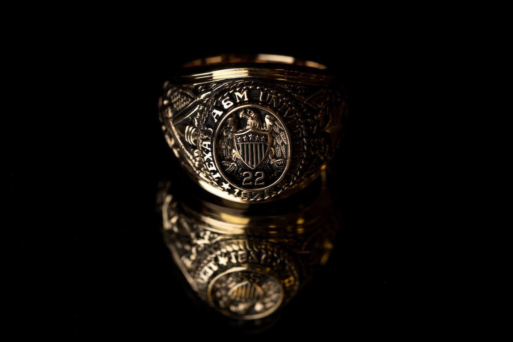 Close up photo of an Aggie Ring against a black background. The image of the ring is reflected on a glass surface.