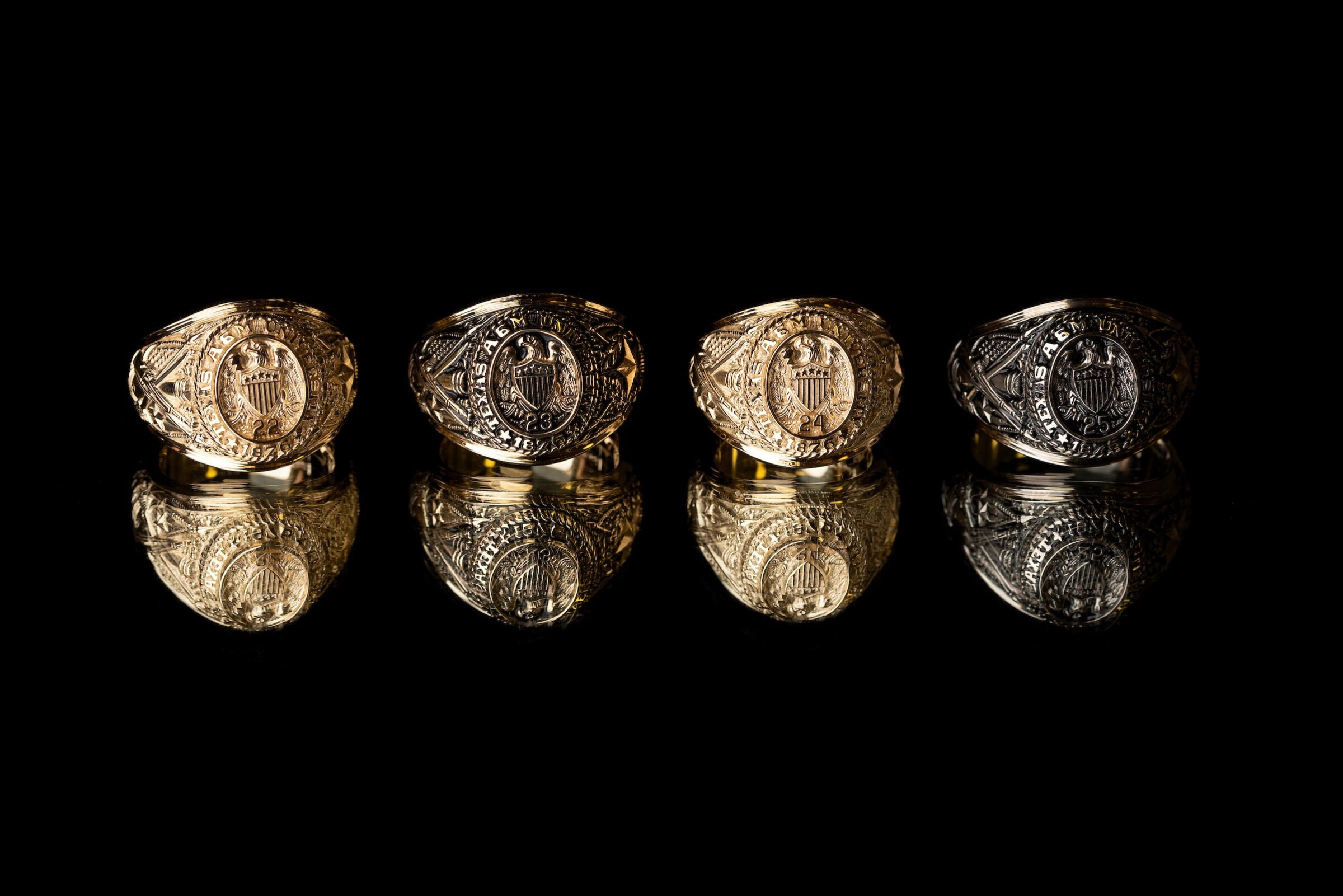 Four Aggie Rings sitting on a reflective surface against a black background.