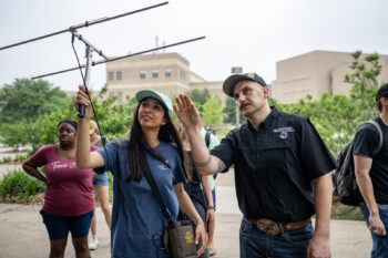 A man in a black shirt (Ty Werdel, Ph.D., assistant professor in the Texas A&M University Department of Rangeland, Wildlife and Fisheries Management) helps a student in a blue shirt and hat try to locate squirrels