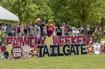A family being photographed in Aggie Park behind a sign that says Family Weekend Tailgate.