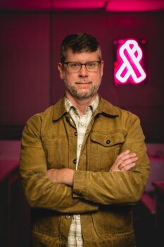 A man wearing a jacket standing with his arms crossed with a neon breast cancer awareness ribbon on the wall behind him.