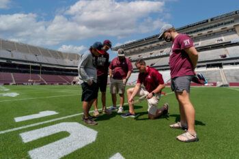 Men gathered around one man who is testing a turfgrass sample inside Kyle Field at Texas A&M.