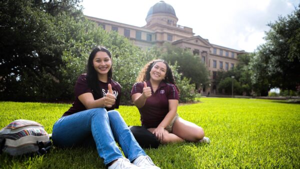 Two Aggie students give thumbs up as they sit on the lawn in front of the Academic Building