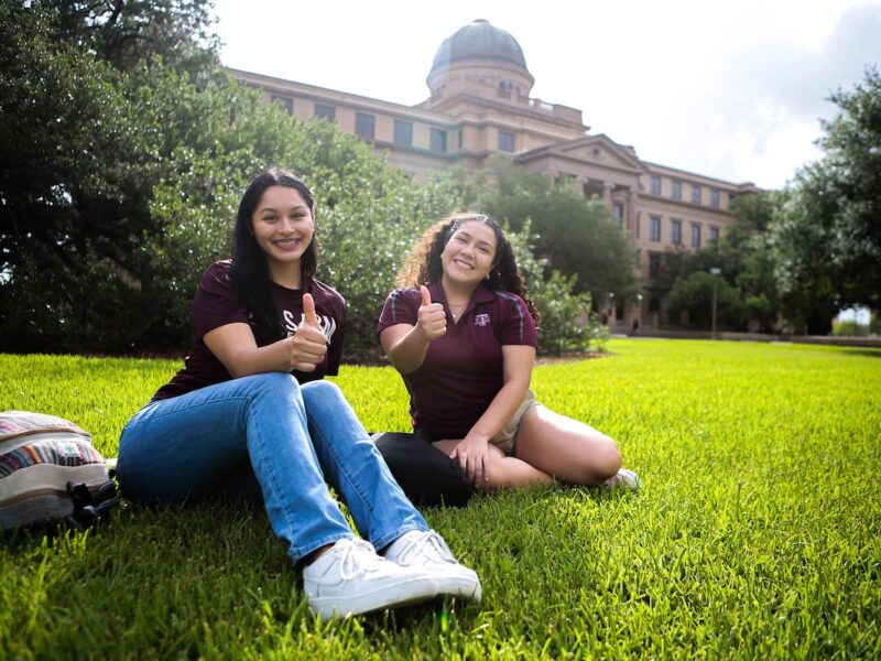 Two Aggie students give thumbs up as they sit on the lawn in front of the Academic Building