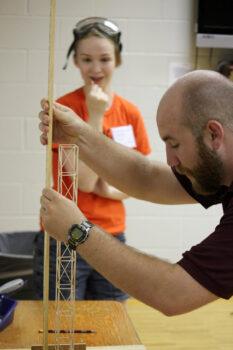 A competitor in a youth science competition watches as a judge measures a tower built out of balsa wood.