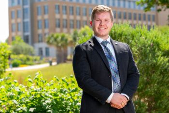 A photo of a young man in a suit and tie standing outdoors smiling