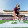 A person in a maroon shirt and cap paints lines on a football field at an empty stadium. He is crouched down, holding a paint sprayer, ensuring the markings are clear and precise.