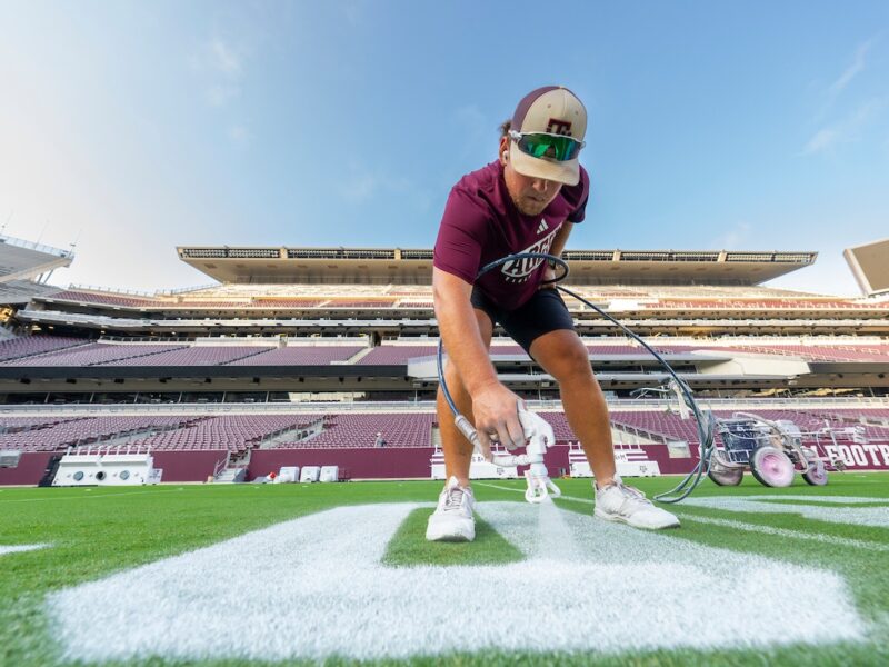 A person in a maroon shirt and cap paints lines on a football field at an empty stadium. He is crouched down, holding a paint sprayer, ensuring the markings are clear and precise.