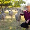 A photo of a man in a cowboy hat kneeling in a fenced pasture with whitetail deer.