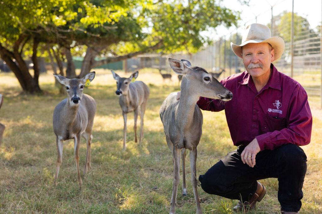 A photo of a man in a cowboy hat kneeling in a fenced pasture with whitetail deer.