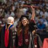 A photo of a graduate holding a diploma tube in the air and smiling to the crowd after crossing the stage during a commencement ceremony.