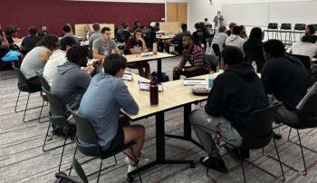 A photo of groups of students sitting at tables in a classroom talking to one person in the center of each table.