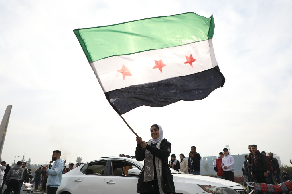 A photo of a Syrian woman waving a flag composed of three horizontal stripes: a green stripe, a white stripe with three red stars, and a black stripe. Others are seen celebrating behind her.