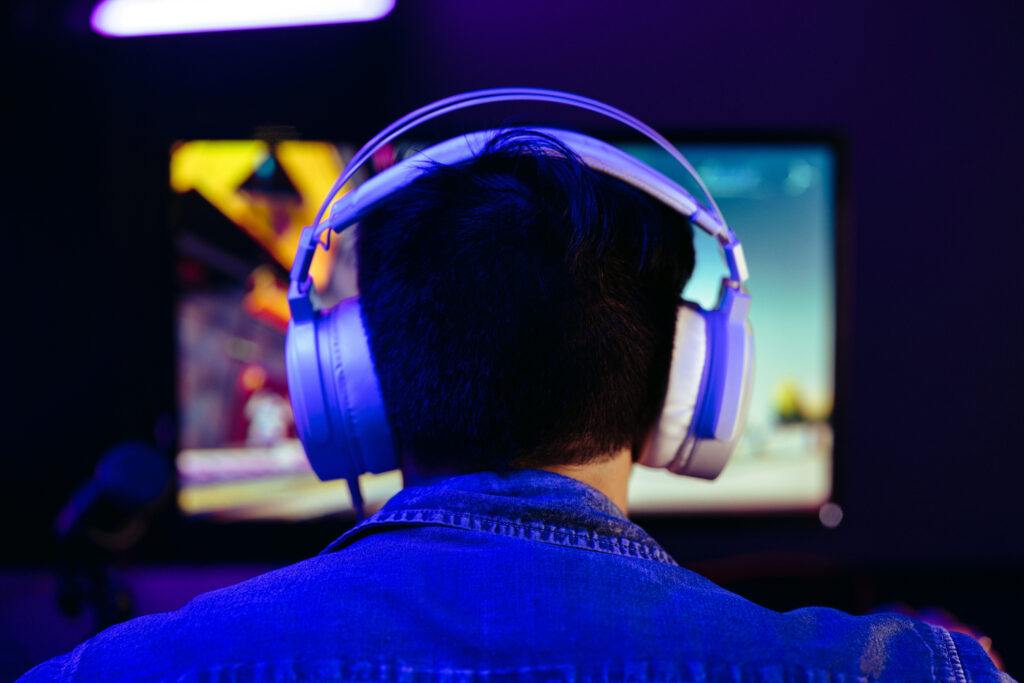 A photo of a man from behind. He is sitting at a desk wearing a large pair of headphones while a monitor glows on the desk in front of him. A microphone sits next to the monitor.