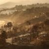 A photo showing a cluster of homes burned to the ground by wildfires. In the background, several large, intact buildings can be seen next to the Pacific Ocean.