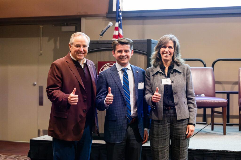 A photo of three people standing in business attire giving the Texas A&M "Gig'em" thumbs-up.