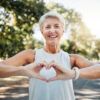 A woman holds her hands together in the shape of a heart while on a walking trail surrounded by trees on a sunny day.