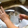 A close-up image of hands filling a glass pitcher with water from a kitchen faucet.