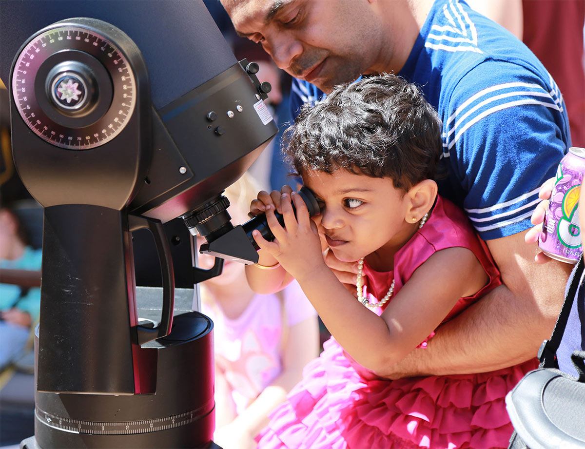a father holds his young daughter up to look through a telescope