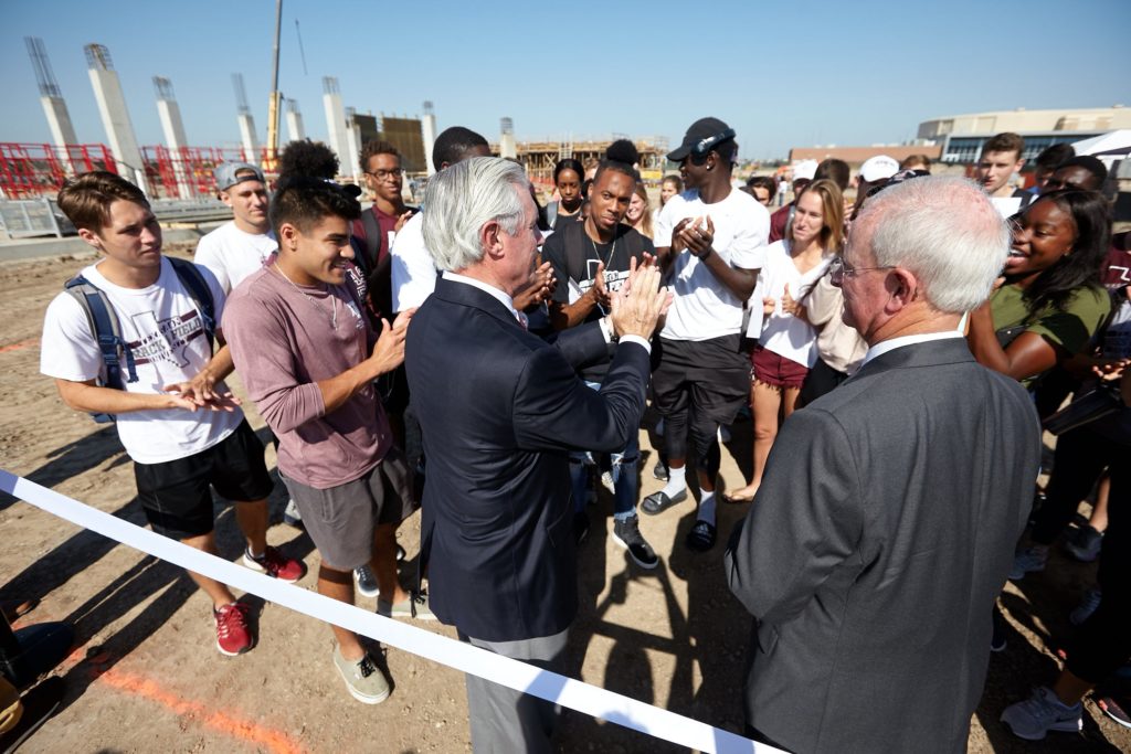 Student-athletes gather around two men in suits at construction site