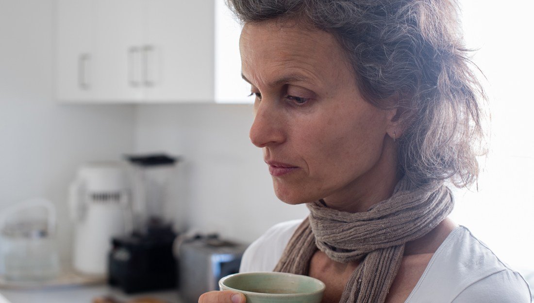 Profile view of middle aged woman with grey hair and scarf holding green cup in kitchen looking worried (selective focus)